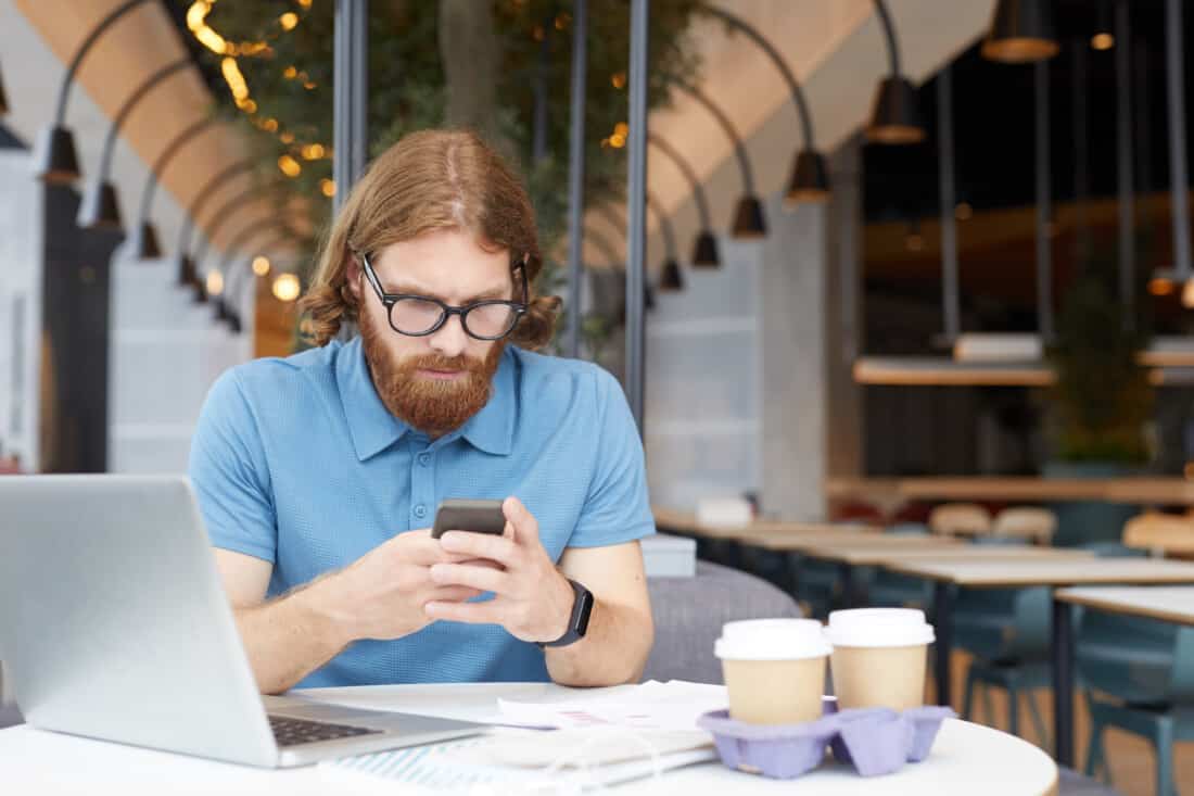 Serious bearded businessman in eyeglasses reading message on mobile phone while sitting at the table with laptop and coffee drinks at the restaurant